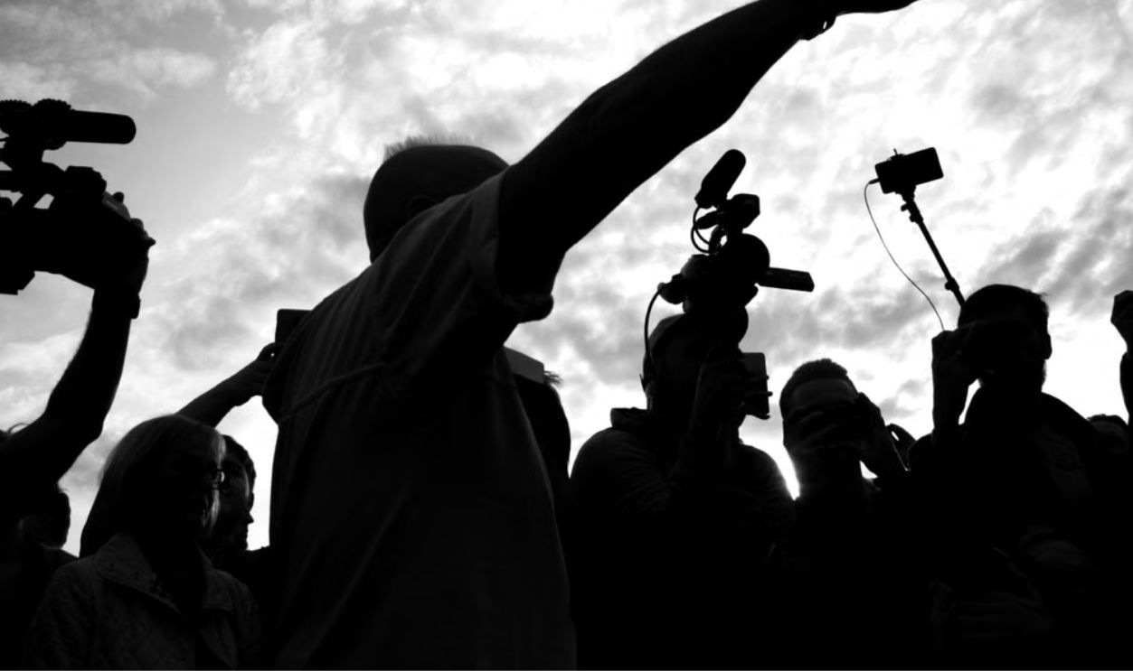 A dramatic silhouette of a media crew with cameras capturing a pointing individual against a cloudy sky
