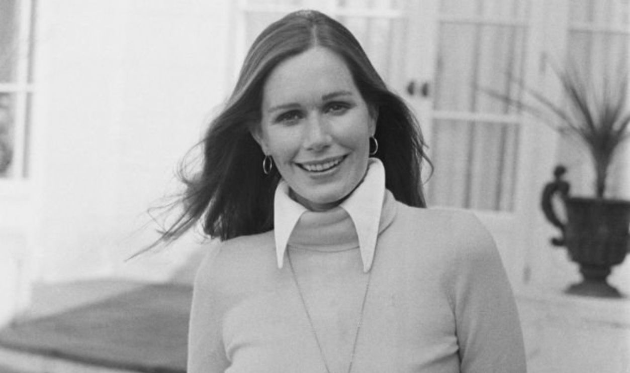 American actress Sally Kellerman wearing a turtleneck sweater with the collar of her blouse protruding over the top, with a locket hanging from a chain around her neck, posing with her hands in her pockets, United Kingdom, March 1973.