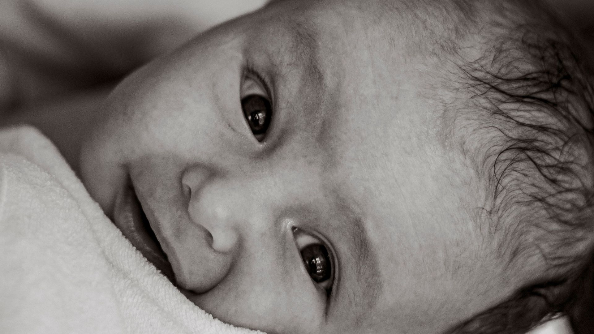 Black and white close-up portrait of a smiling newborn wrapped in a robe with a cross pattern.