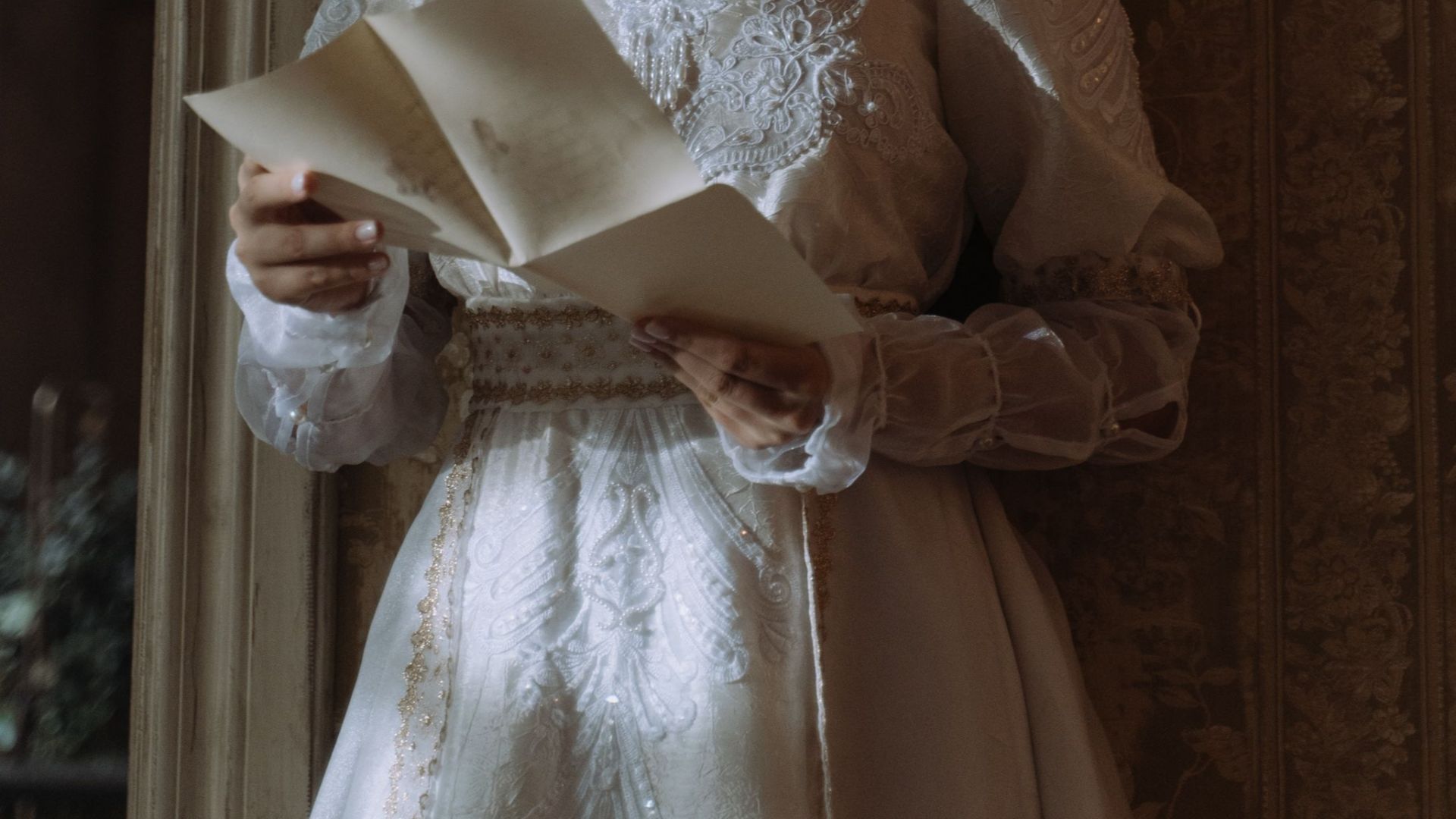 Woman in white dress reading a letter by a vintage frame indoors.