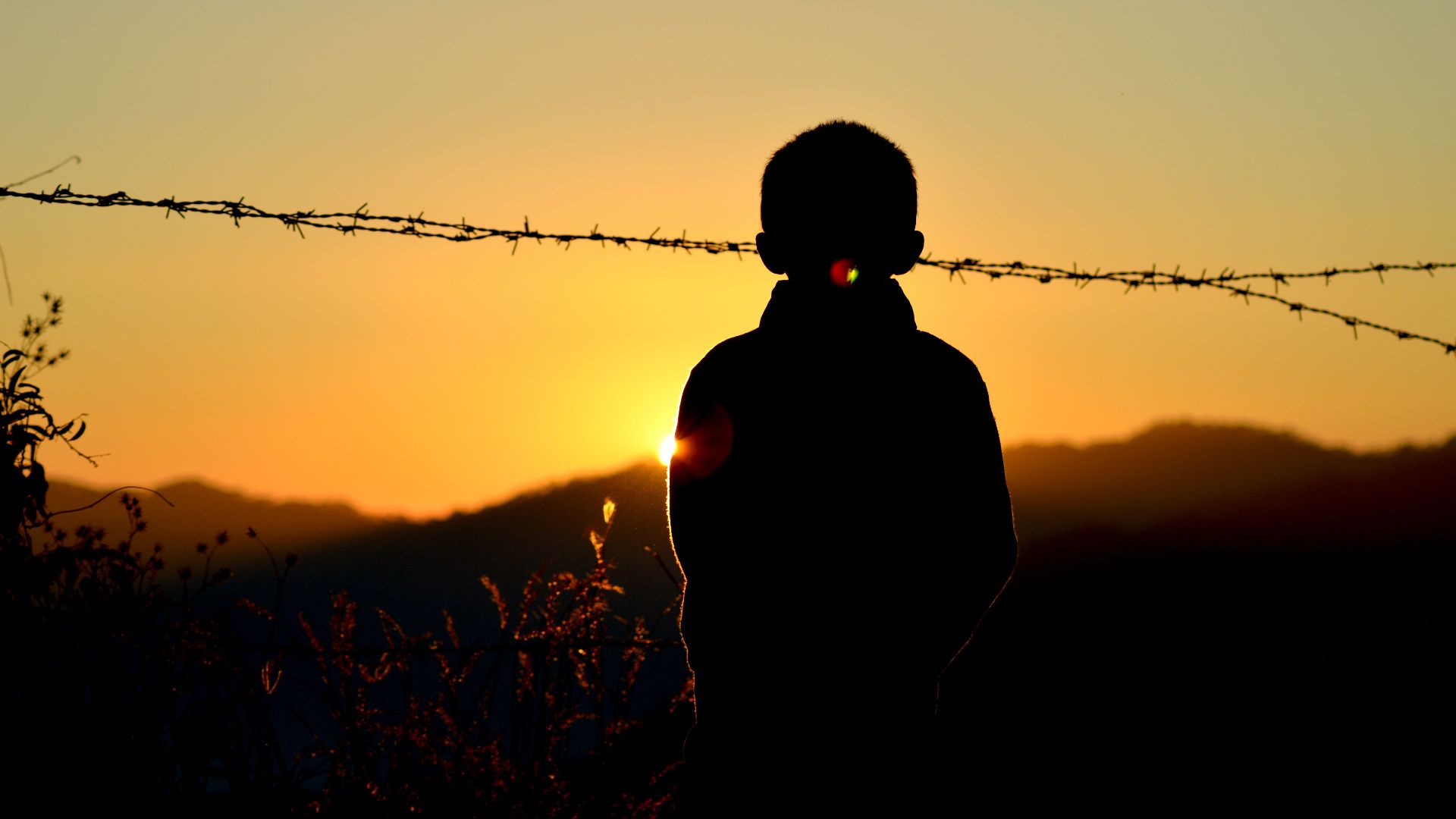 A child's silhouette against a sunset backdrop with mountains and barbed wire.
