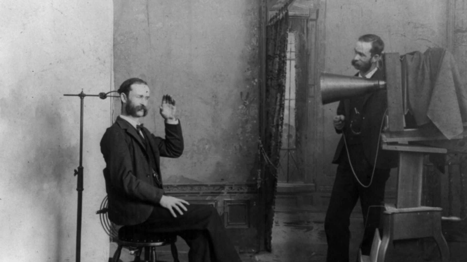 TITLE: A photographer appears to be photographing himself in a photographic studio: Wheeler, Berlin, Wis.
A composite photograph showing a photographic studio interior. One man is seated on a stool near an adjustable clamp to hold his head steady during a
