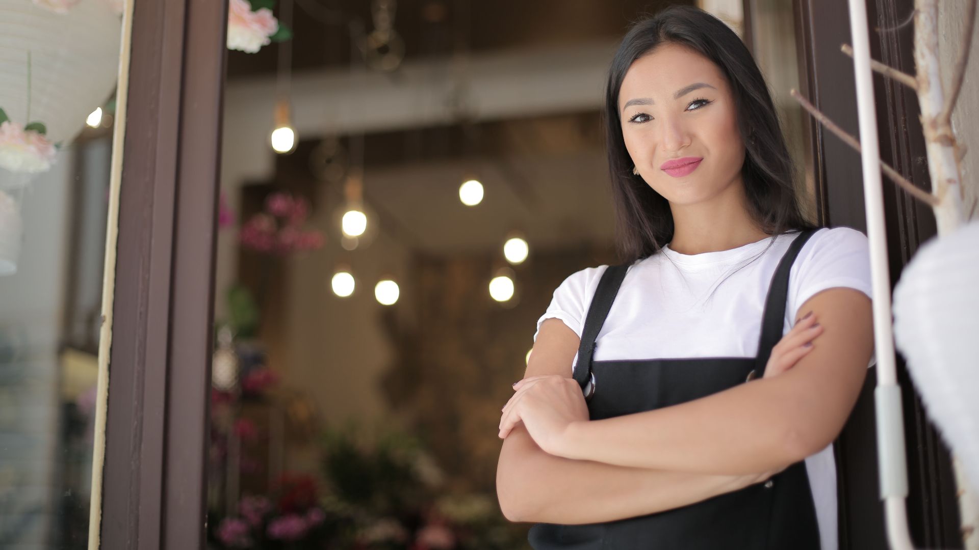 From below of young confident Asian female standing with crossed arms at entrance of floral shop and smiling at camera