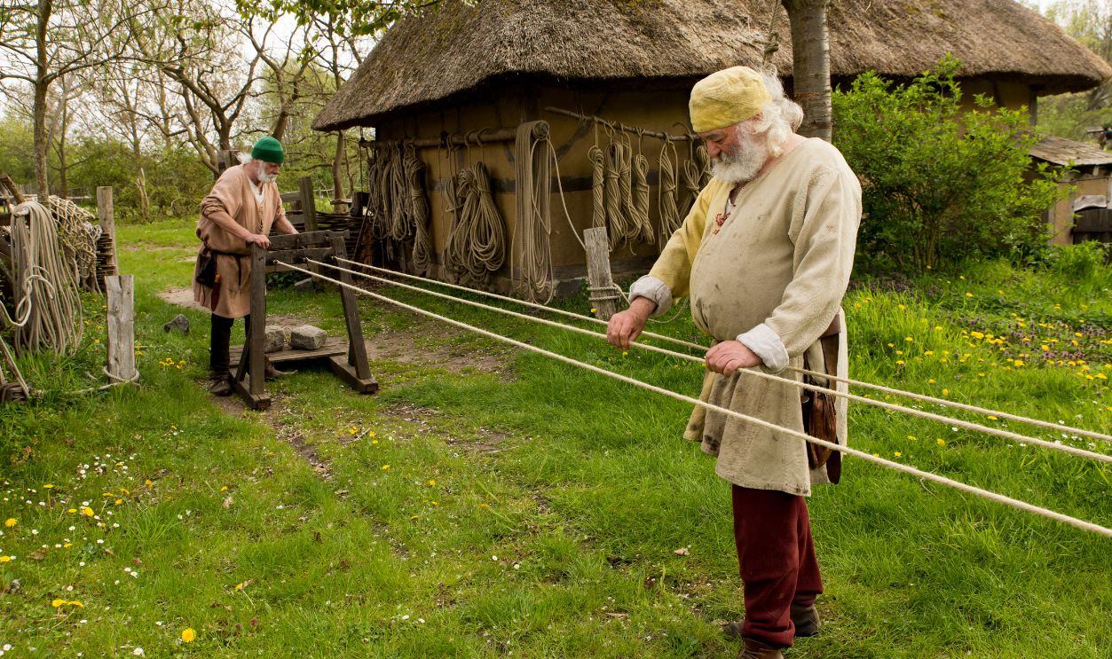 NYKOBING FALSTER MAY 01: Two men in traditional cloth from the middle ages produce rope at the outdoor Medieval Centre on May 01, 2019 in Nykobing Falster, Denmark. The Medieval Centre is built around the village Sundkobing at a fjord as it was around the year 1400. Houses are authentically designed and built with the people who populate the village, men, women, children, peasants, craftsmen, warriors and knights etc dressing and work as they would back in 1400. Canons and large trebuchets can be seen firing missiles with knights in armour on horseback competing with lances and swords, dyers colour textiles with plants and in the convent garden nuns grow medicinal herbs. 