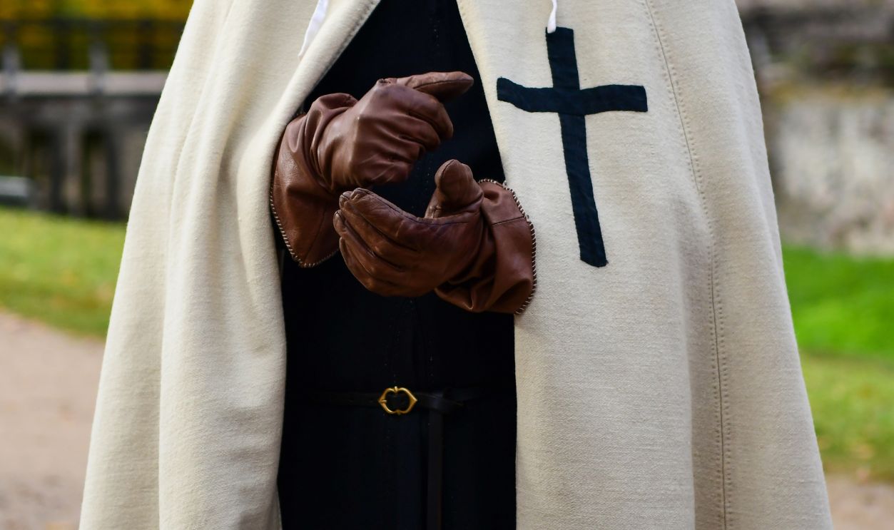 Man in black clothes and beige cloak with black cross. Wearing old gloves, standing outdoors near a castle in autumn. Historical, mystical, medieval atmosphere.