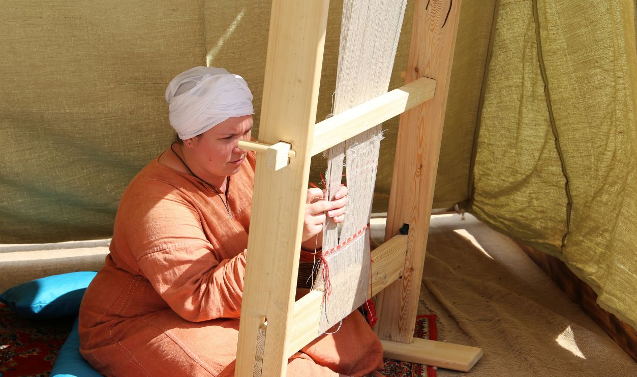 Moscow, Russia - June 2019: Woman in medieval clothes sitting near the wooden loom during the Moscow historical festival Times and epochs. Female weaver at work