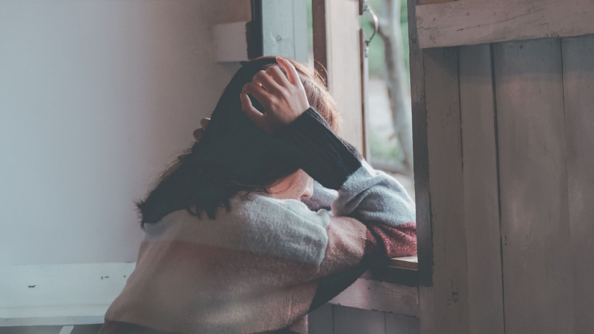 A woman sits indoors gazing thoughtfully out of a window, evoking emotions of loneliness and reflection.