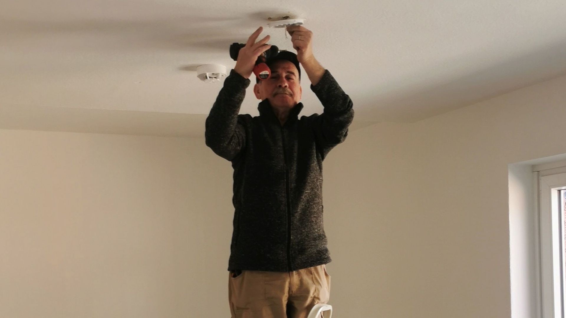 Adult man on ladder fixing a ceiling light in an empty room with beige walls.