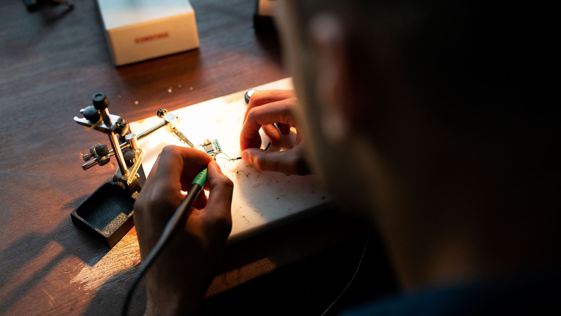 A focused engineer soldering a circuit board under bright light in a workshop setting.
