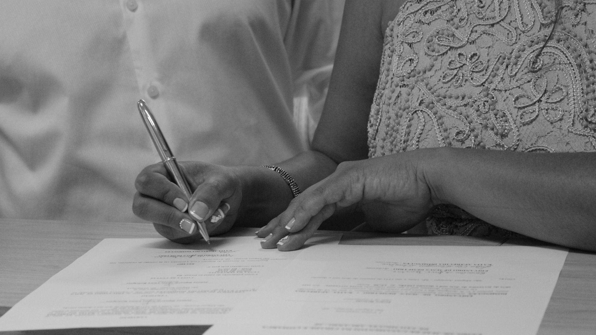 Black and white image of a couple signing documents, focusing on their hands and pen.