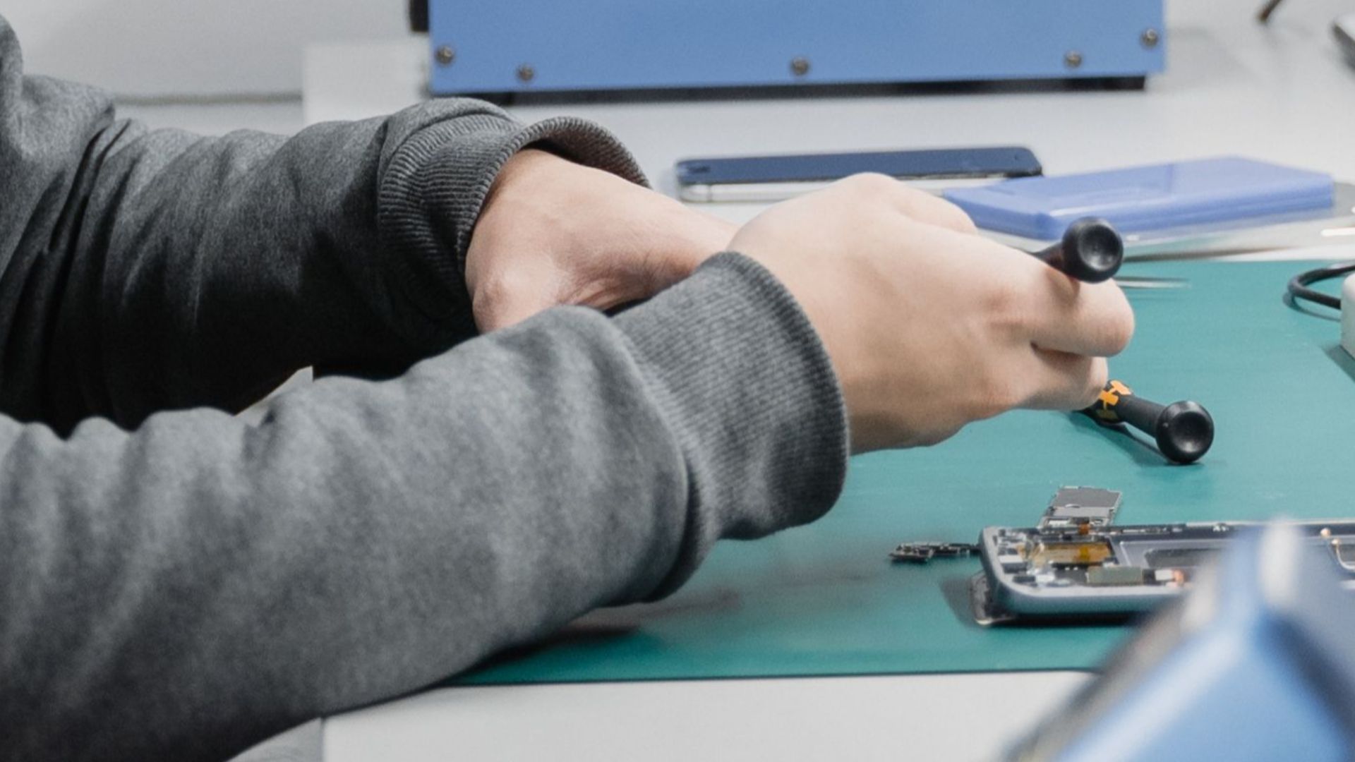 A young male engineer repairs electronics at a workshop desk with tools and equipment.