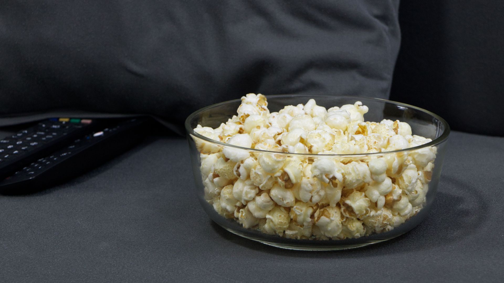 Glass bowl filled with popcorn next to TV remotes on a gray sofa. Perfect for cozy movie nights.
