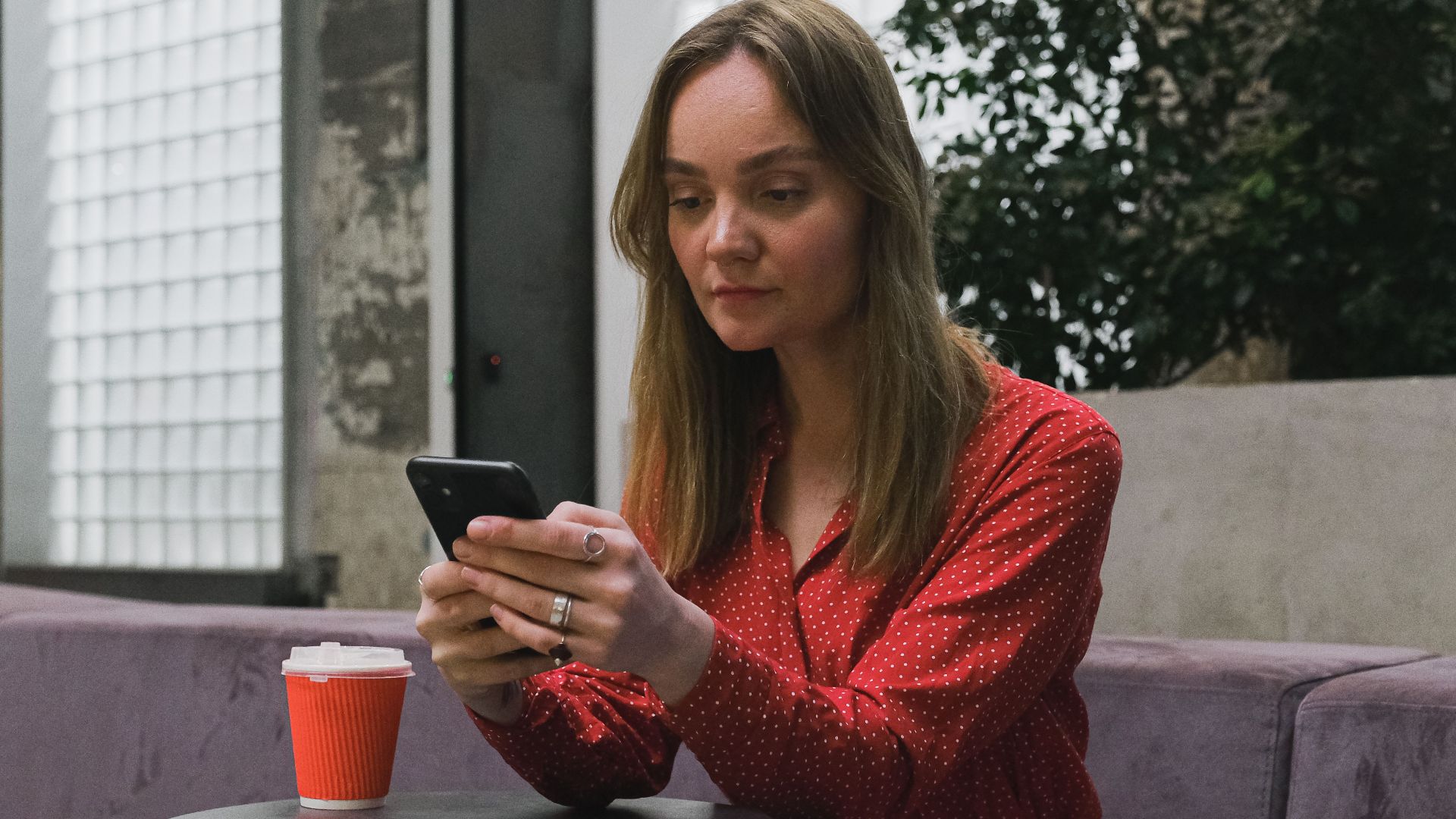 Young woman in red dress sitting indoors, using smartphone at a table with a cup of coffee.