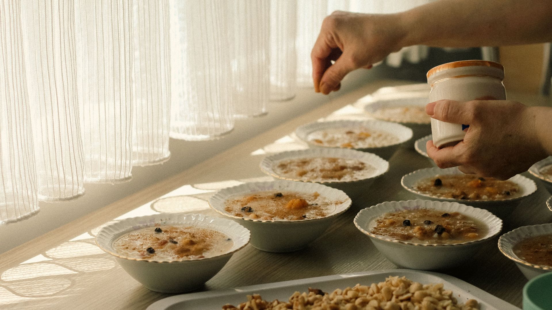 Person adding toppings to bowls of oatmeal on a sunlit table, featuring nuts and raisins.