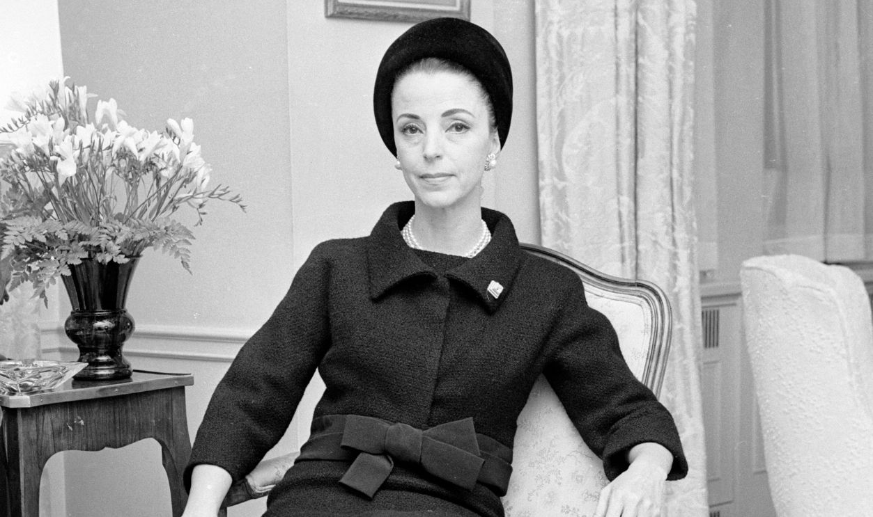  Gloria Guinness seated in her suite in the Waldorf Towers wearing a black wool Givenchy jacket bordered in black faille over a sleeveless dress with a black Balenciaga hat and diamond ring and lapel pin as well as pearls and diamond and pearl earrings; black round-toe pumps completed the dinner outfit (Guinness owned the exact same outifts in mauve and blue linen with a beige hat