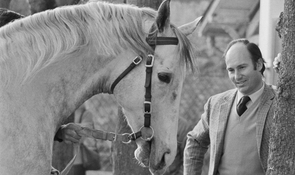 Aga Khan IV, wearing a tweed jacket over a sweater with a shirt and tie, as he stands with his horse Zeddaan, United Kingdom, 18th March 1973