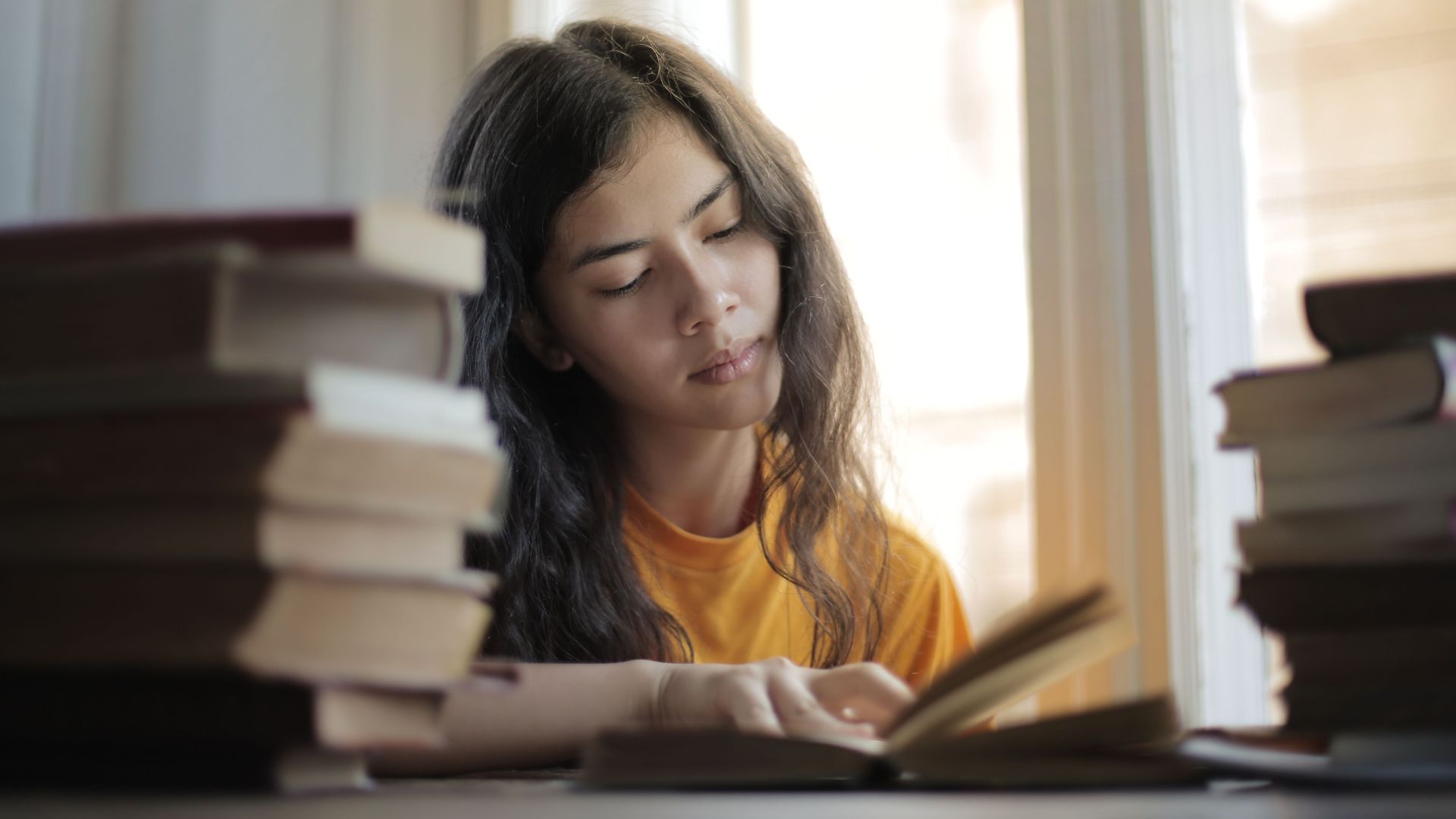 Young smart Asian woman reading book while sitting at table by window in soft daylight