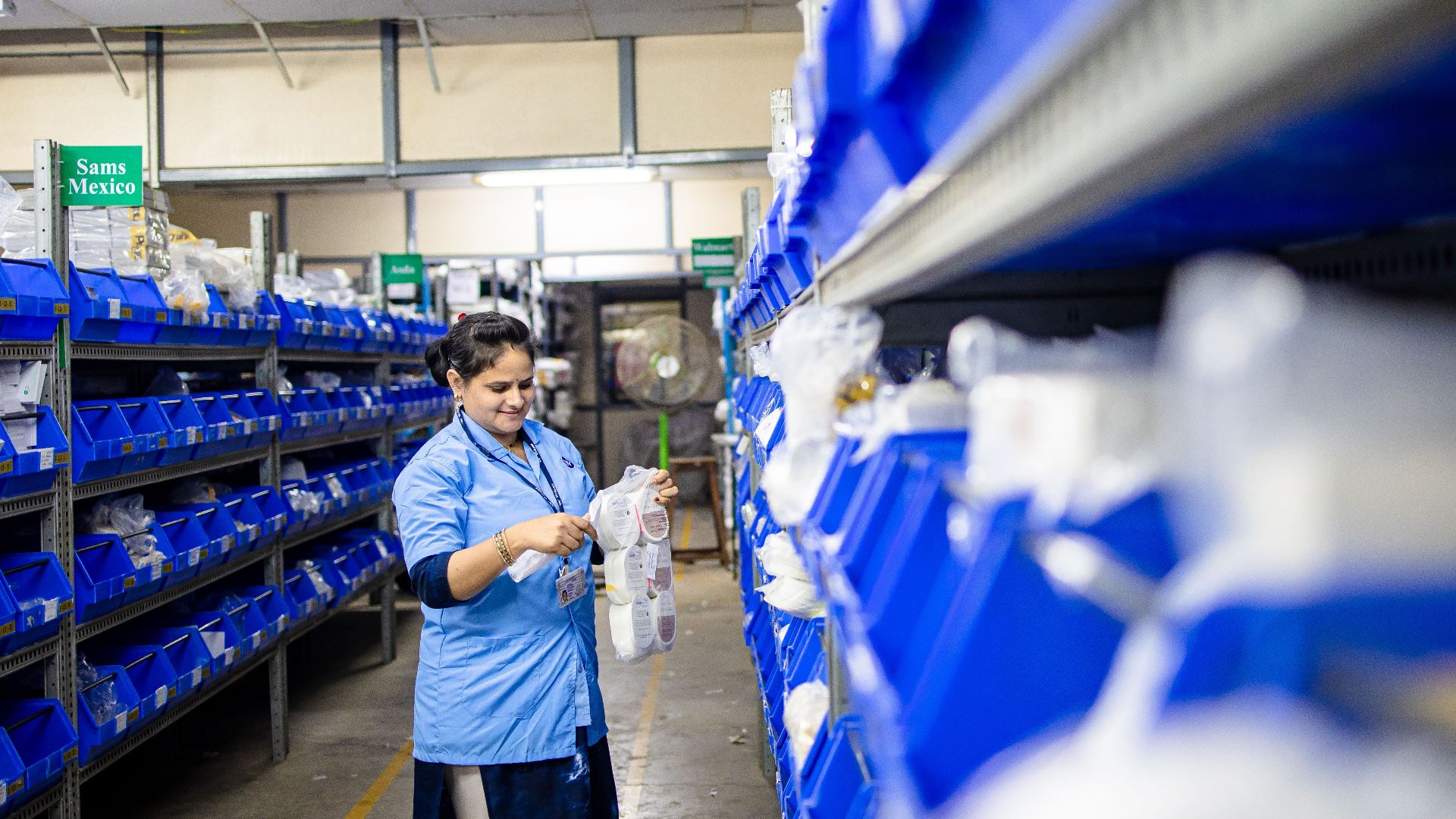 Female worker organizes shelves in a warehouse. Industrial setting with labeled storage bins.