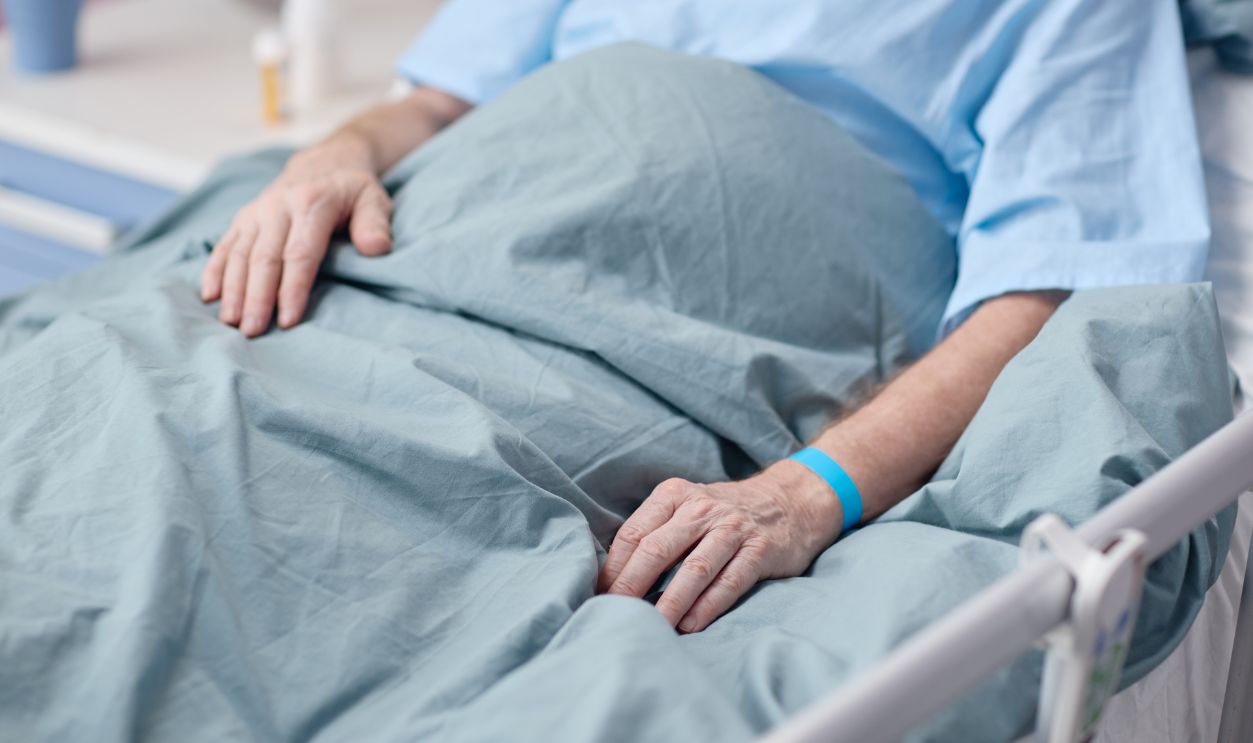 Senior Caucasian man lying in hospital bed with medical wristband visible, resting hands on blanket in vascular center, showing partial body and hospital environment