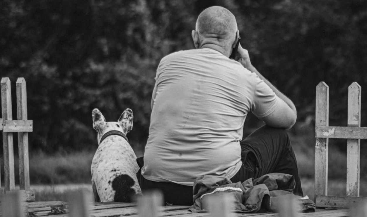 A man and his dog sitting outdoors on a wooden terrace, captured in black and white