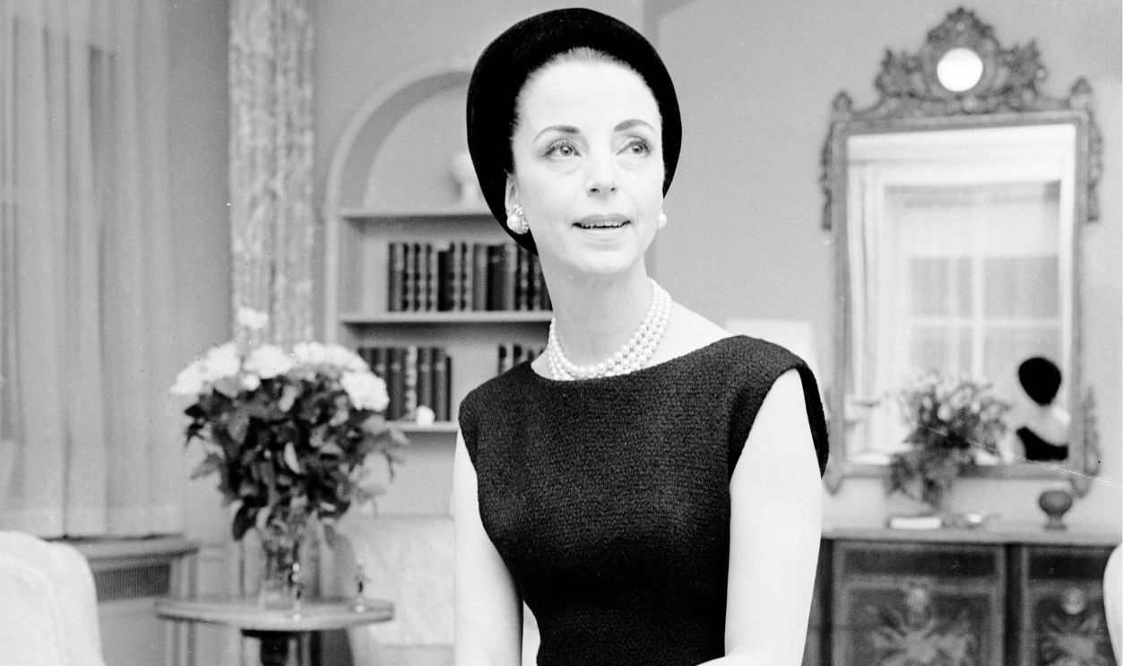 Gloria Guinness seated in her suite in the Waldorf Towers wearing a black wool Givenchy jacket bordered in black faille over a sleeveless dress with a black Balenciaga hat and diamond ring and lapel pin as well as pearls and diamond and pearl earrings; black round-toe pumps completed the dinner outfit (Guinness owned the exact same outifts in mauve and blue linen with a beige hat)