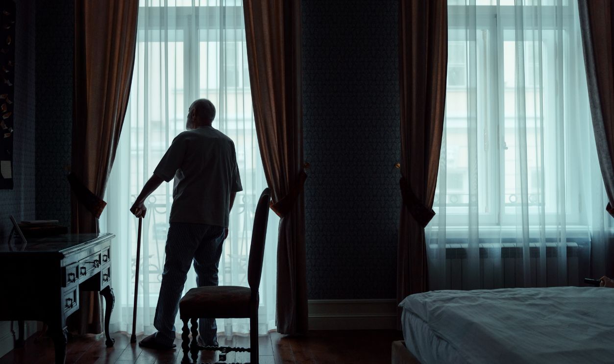 Silhouette of an elderly man standing with cane, gazing out of curtain-covered window