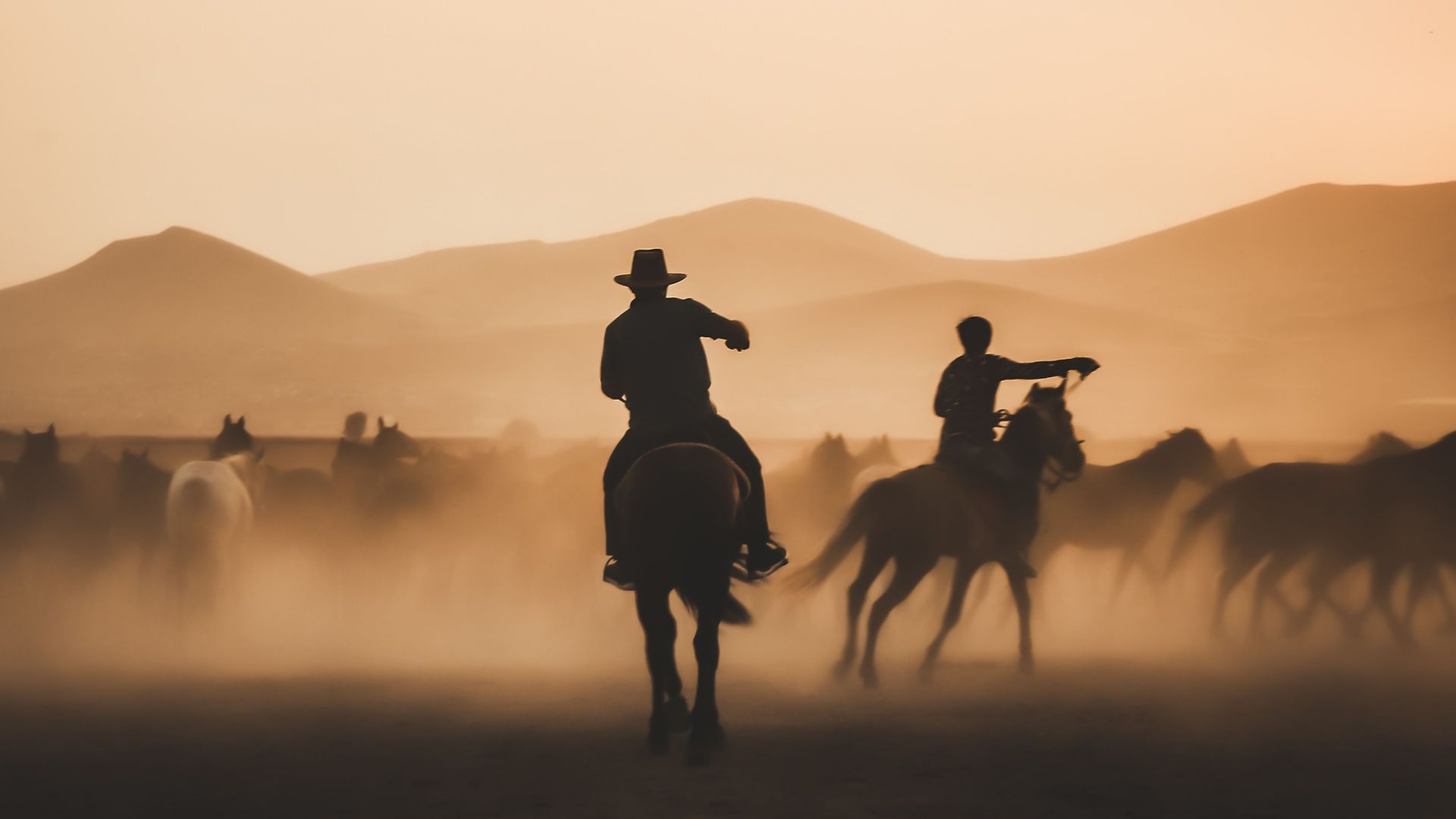 Silhouetted cowboys herding horses across a dusty landscape during a stunning sunset.