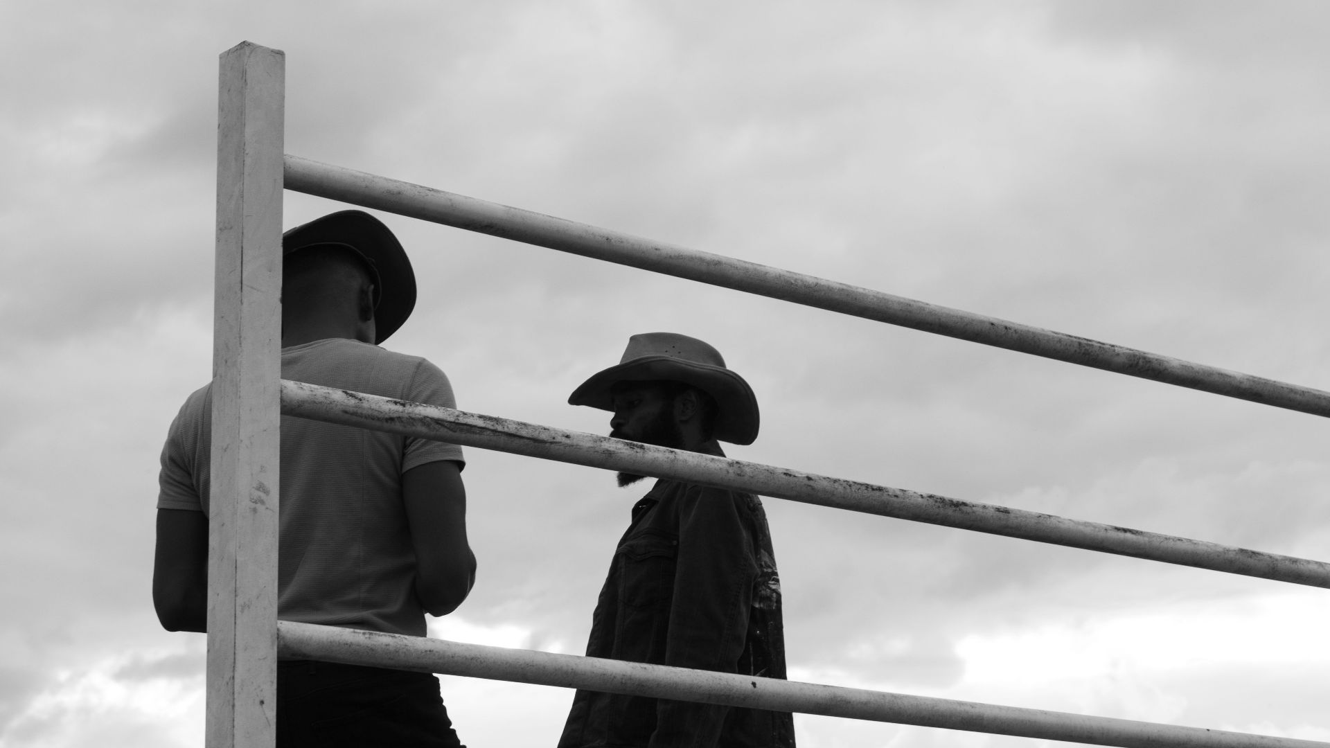 Two cowboy silhouettes in hats and boots leaning on a fence at a ranch.