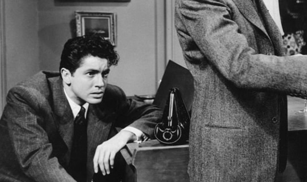 Farley Granger watches a situation unfold while seated at a piano, as James Stewart holds a gun to John Dall's chest in Alfred Hitchcock's 1948 thriller, Rope. 