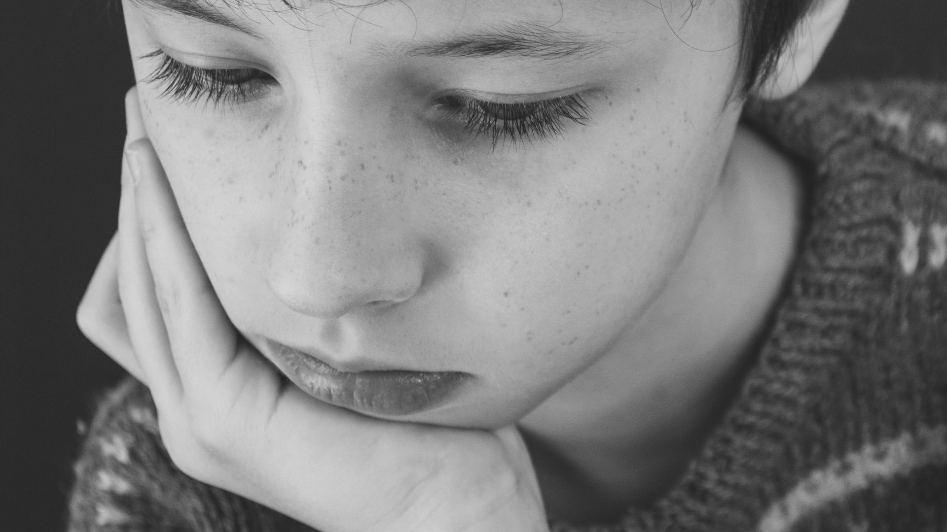 Black and white of sad boy with short dark hair in casual clothes covering face with hand while looking down