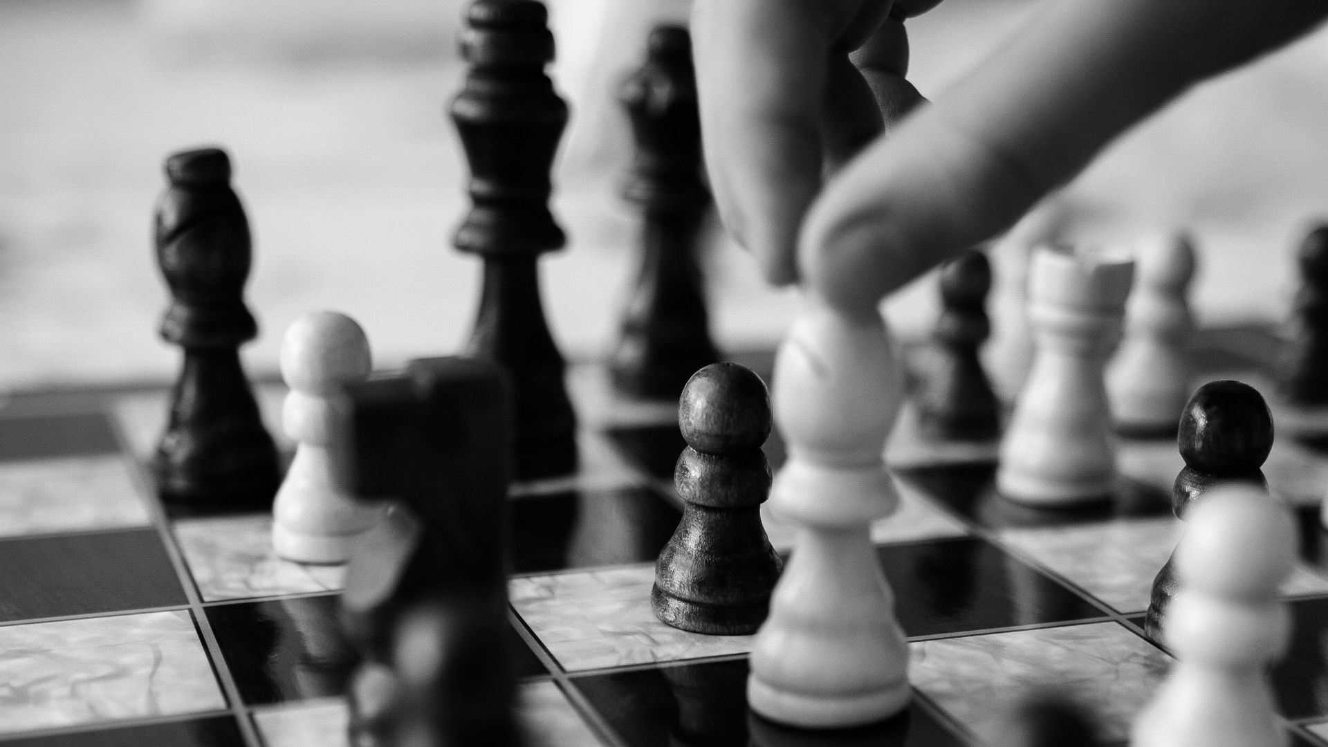 Black and white image of a hand moving chess pieces on a board, highlighting strategic play.