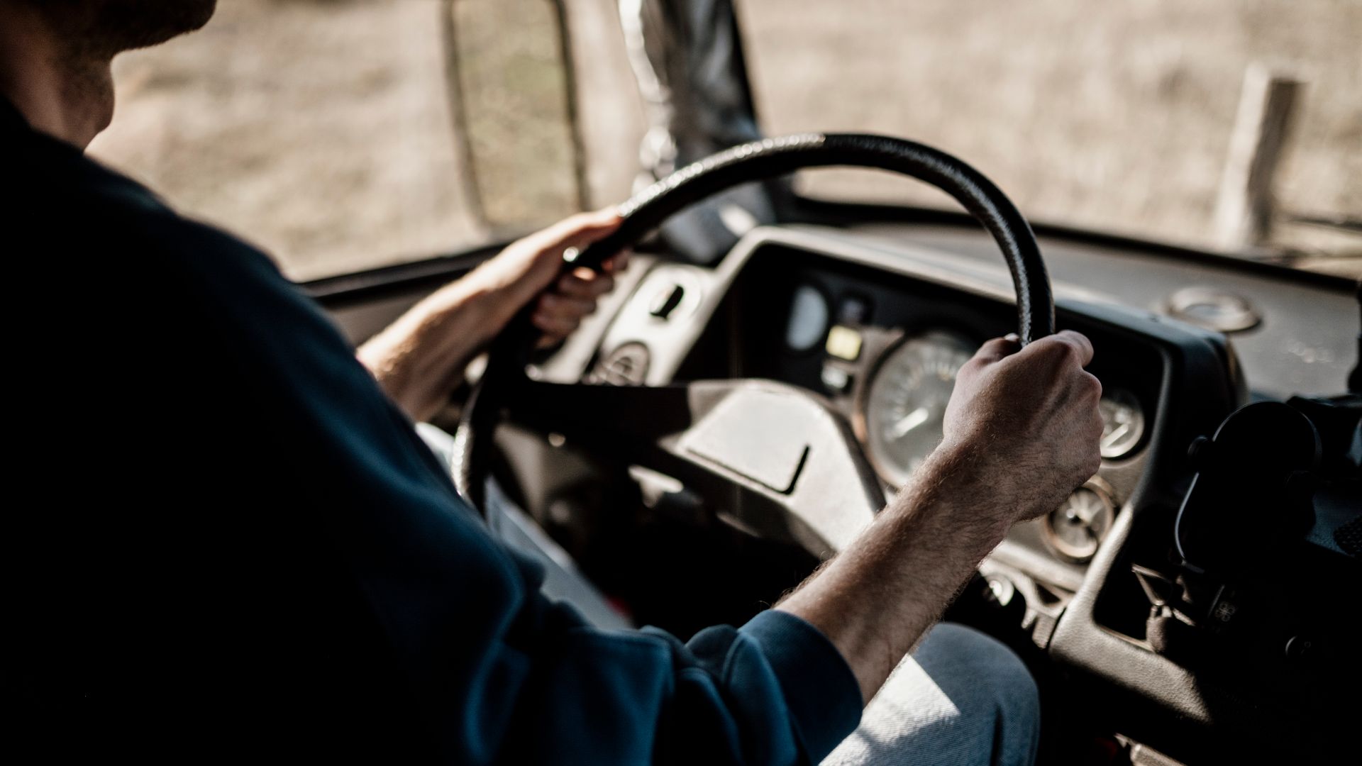 A person steers a truck with both hands on the wheel. The focus is on the driver's hands and the dashboard as fields stretch out ahead. Sunlight fills the cabin during the drive.