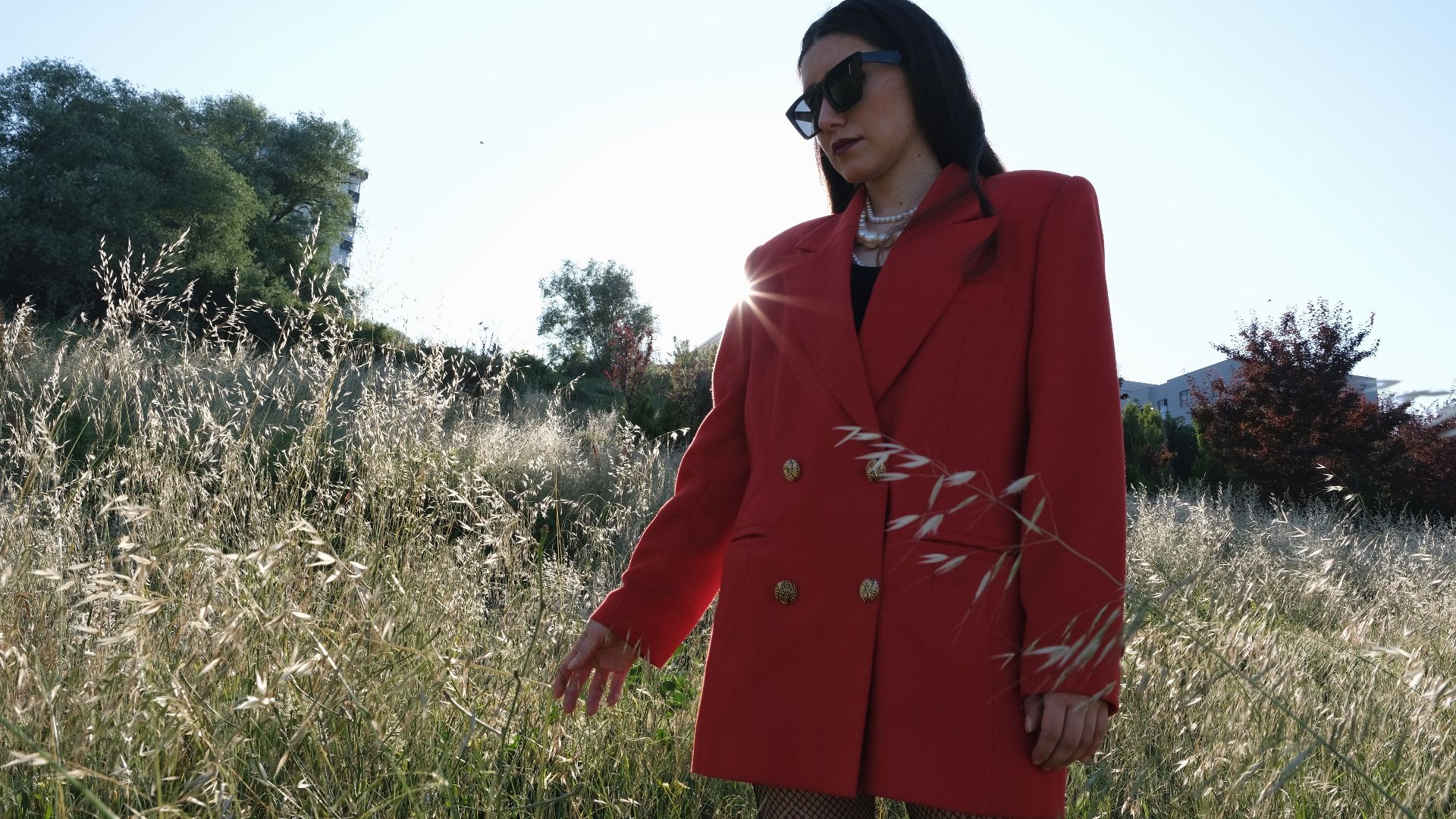 Fashionable woman wearing a red coat stands in a sunlit meadow, exuding elegance.