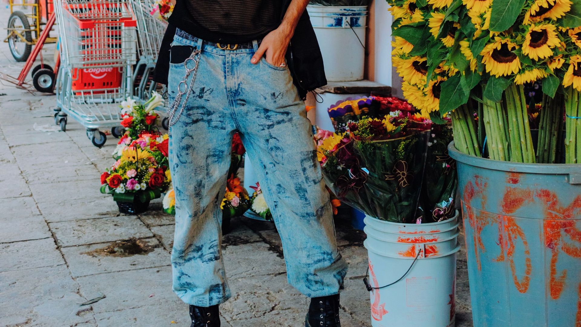 A fashionable man in sunglasses standing by a vibrant sunflower display on an urban sidewalk.