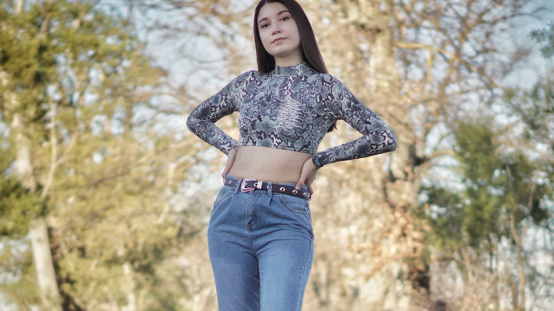 Woman in casual crop top and denim jeans posing in a natural outdoor setting.