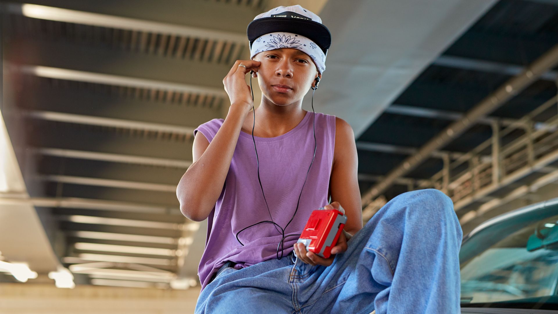Teen boy in casual attire enjoying music with a Walkman under a bridge.