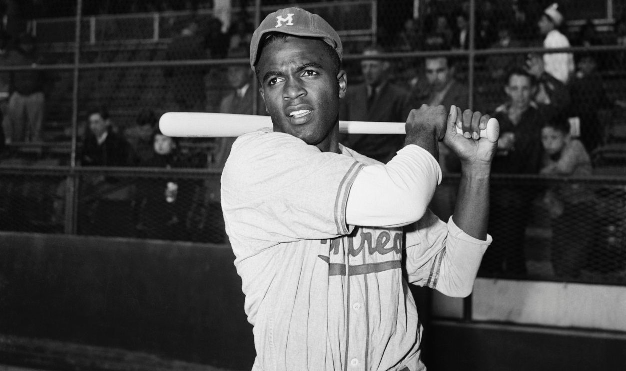 Jackie Robinson, first Black man to be signed up by a Major League baseball team, is shown in post-swing position in front of the stands. Robinson changed uniforms after playing with Montreal in an exhibition game against the Dodgers.