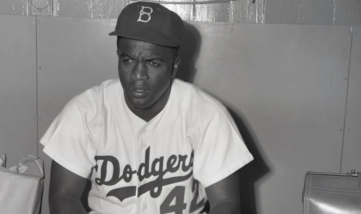 Brooklyn Dodgers baseball player #42, Jackie Robinson is shown sitting in uniform holding baseball and glove.