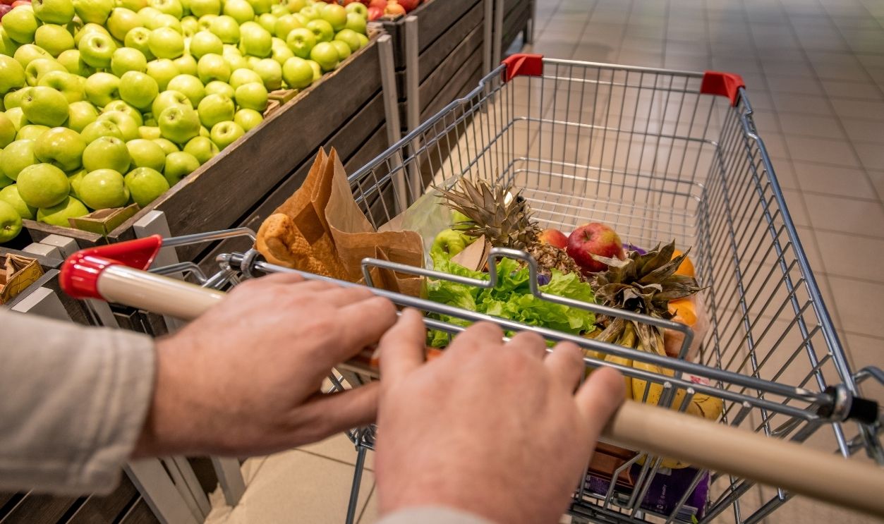 Hands of aged male customer pushing shopping cart with food products while moving along fruit display with fresh apples in supermarket