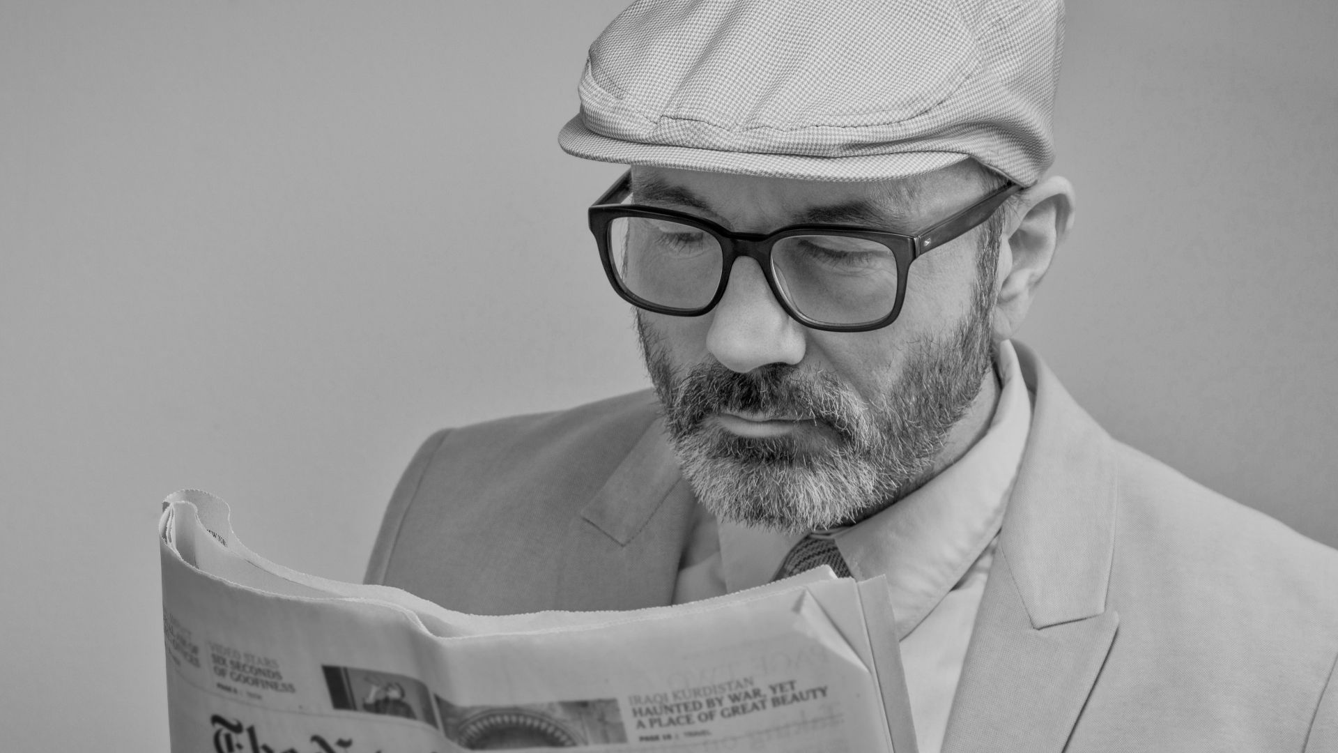 A man in a suit, wearing glasses and a cap, reads The New York Times.