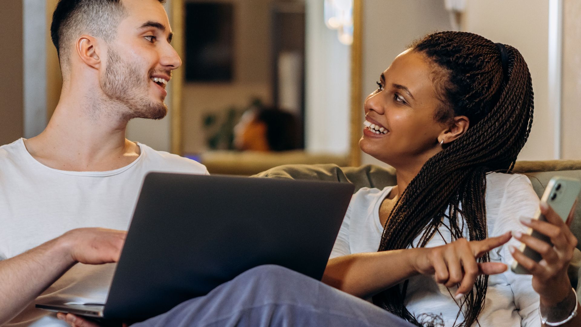 Interracial couple bonding over technology, sitting on a couch indoors with a laptop and smartphone in hand.