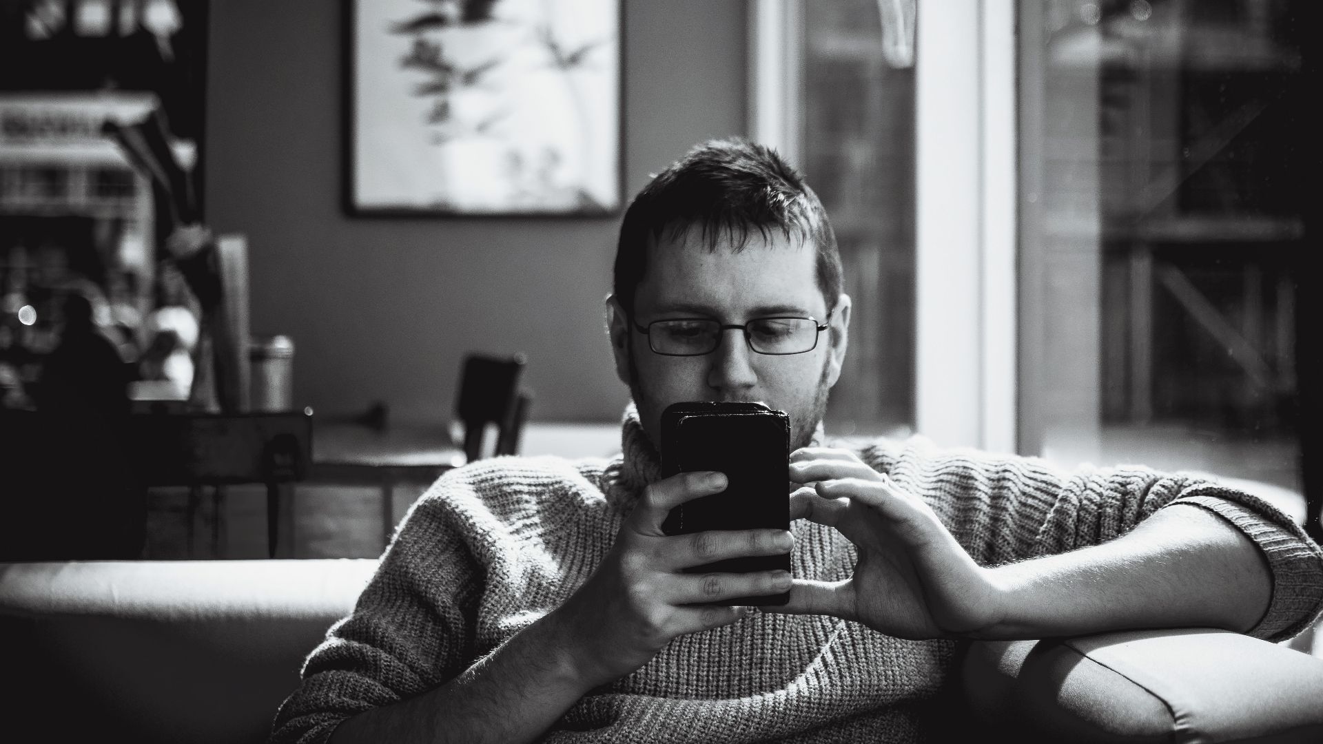 A man in a cozy indoor space engrossed with his smartphone, sipping coffee.