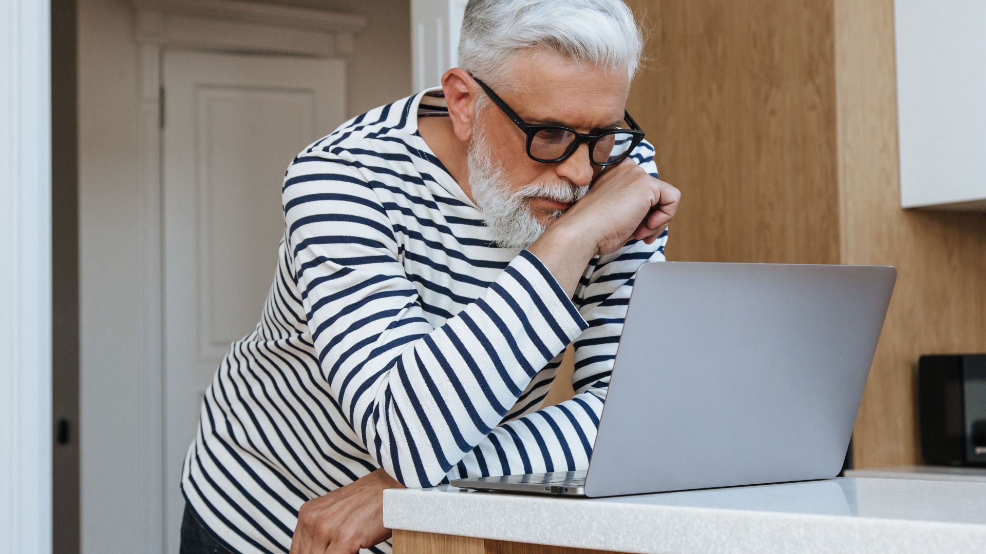 A senior adult man with glasses concentrating on his laptop at home, symbolizing modern working environments.