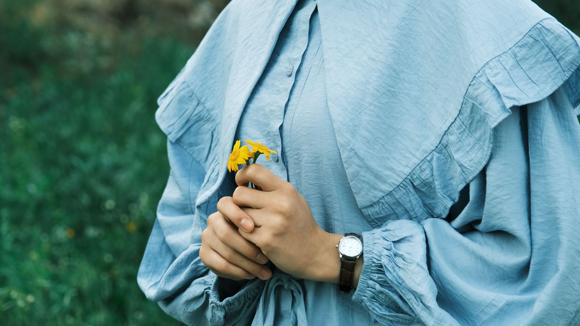 A woman in a blue dress holds a yellow flower in a green outdoor setting, capturing a serene moment.