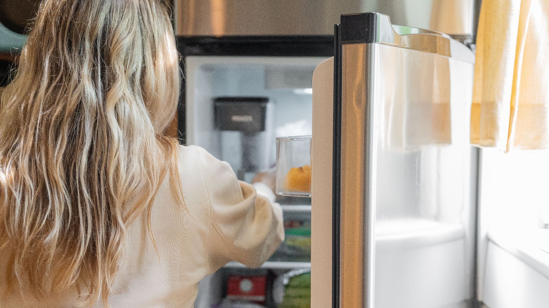 Back view of a blonde woman retrieving food from a refrigerator in a bright kitchen.