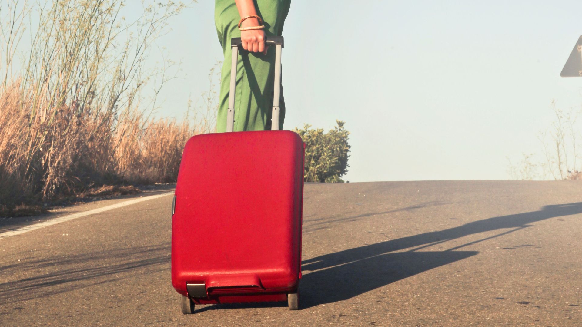 Fashionable woman in green attire with red suitcase traveling on sunny road.