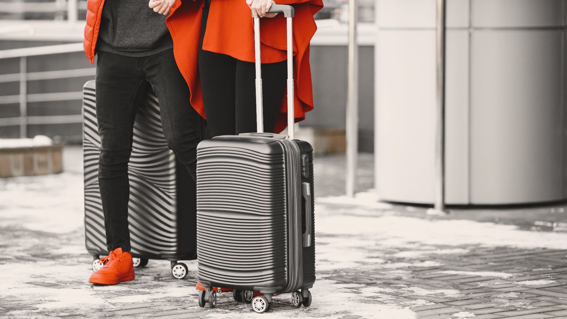 A fashionable couple in winter attire with sleek luggage, standing outdoors on a snowy day.