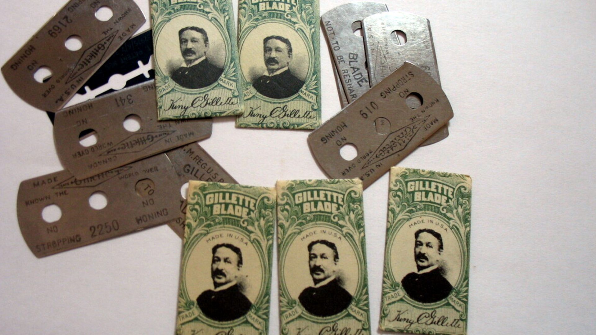 Original-style Gillette safety razor blade wrappers and blades. Most of the blades are of the three-hole design. One (partially obscured by the others) is likely of the more modern used from 1933 until today. Possibly a Gillette Blue blade as it's dark in
