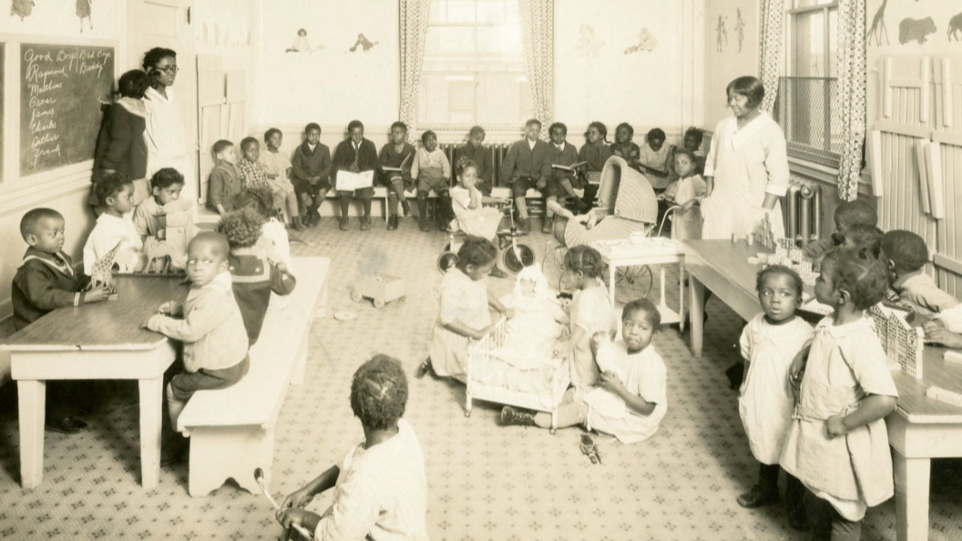 Depicts two African American women teachers overseeing a class of African American grade school children. The younger children play on the floor and at tables with blocks. The older ones sit and read on benches lining the wall. On the floor, a group of gi