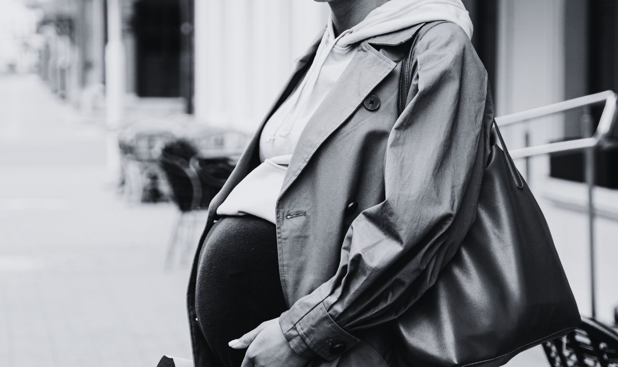 A smiling pregnant woman in a brown coat holding a bouquet
